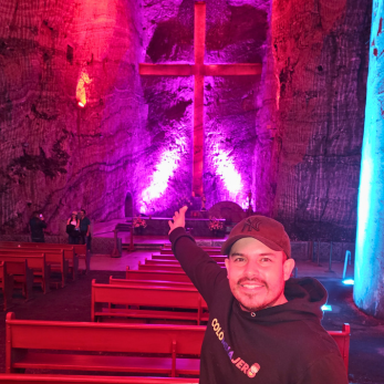 Hombre con gorra y buzo negro señalando la gran cruz iluminada de color púrpura dentro de la Catedral de Sal de Zipaquirá, con las bancas de la iglesia y paredes de roca salina al fondo.