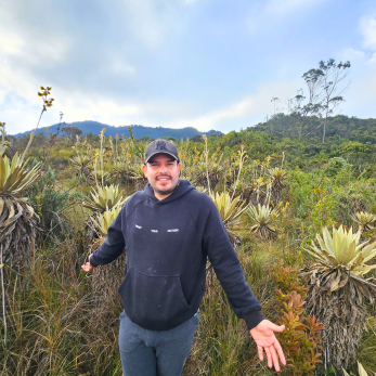 Hombre joven con gorra y buzo negro posando en un campo de frailejones en el Santuario Las Moyas, Bogotá, con montañas verdes de fondo bajo un cielo nublado.